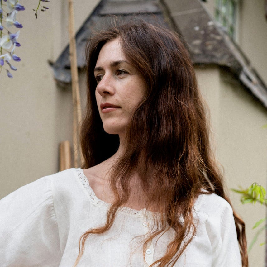 Woman in a white outfit standing in a garden with greenery and a rustic gate.